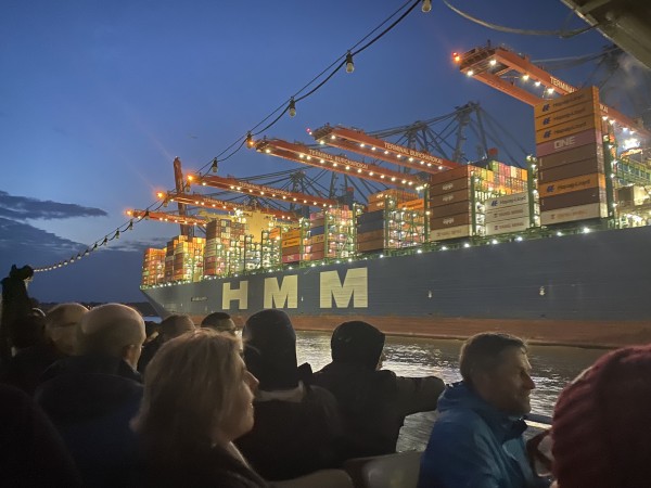 CiviCamp Hamburg 2024 Teilnehmende bei der nächtlichen Hafentour im Vordergrund mit einem großen Containerschiff im Hintergrund